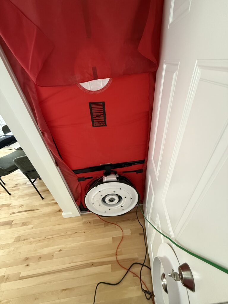 Close-up of a red blower door shroud and fan in a doorway beside a white interior door on a hardwood floor - blower door testing in Ashfield, MA