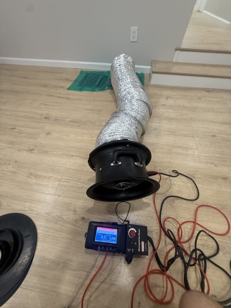 Duct Blaster fan on a hardwood floor with hose run to a sealed return grille by the stairs – HEEC duct leakage testing for Adams, MA homes