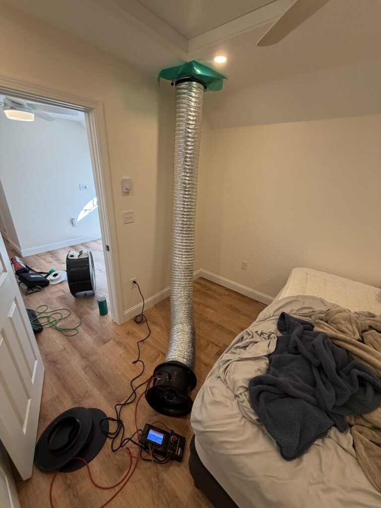 Duct Blaster hose sealed to a ceiling return grille in a bedroom next to an unmade bed - duct leakage testing in Chicopee, MA