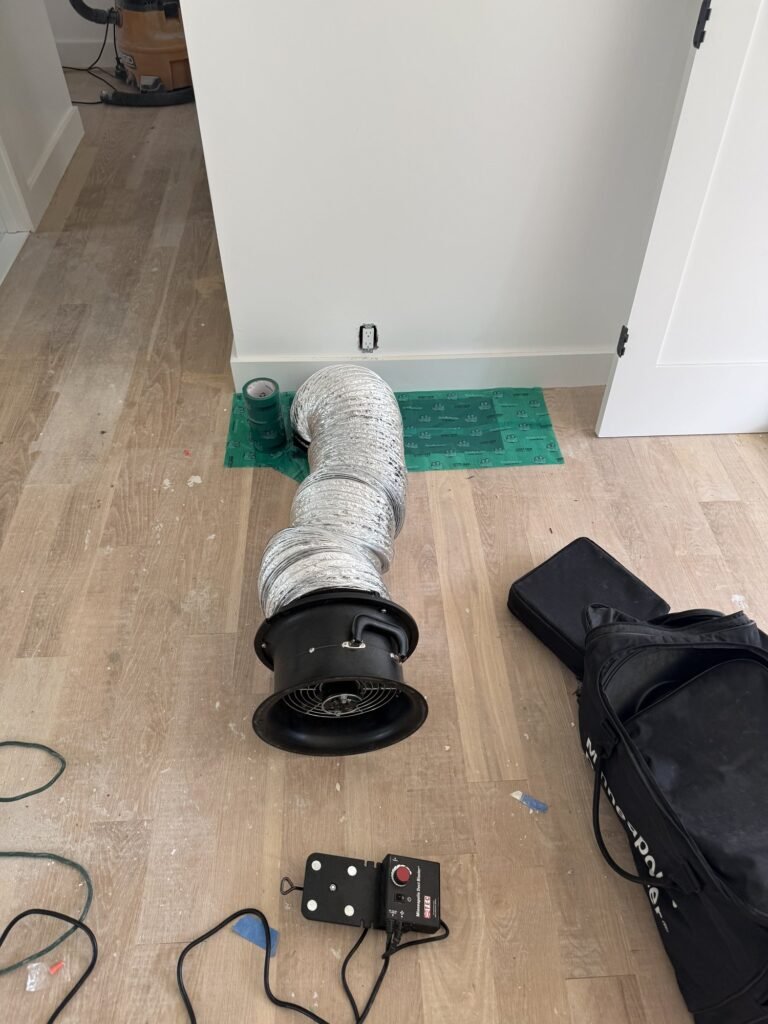 Flex duct from a taped low return grille to the Duct Blaster fan on a light oak floor - duct leakage testing in Acton, MA