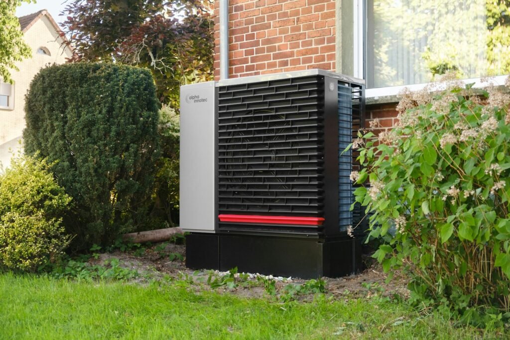 Air-source heat pump outdoor unit set among garden shrubs beside a brick home - HERS rating in Groton, MA